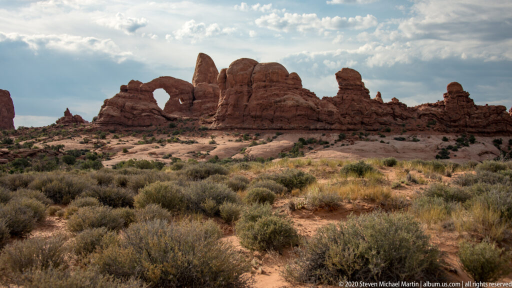 Panoramic line of sandstone arches and pinnacles above a scrub-filled basin and faint footpath.