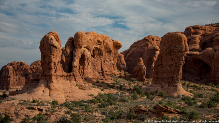 Cluster of red sandstone towers and alcoves with shrubs growing in the gaps between formations.