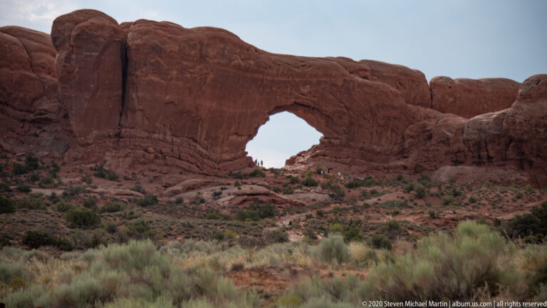 Large sandstone arch above a sloping path, with small visitors beneath the opening.