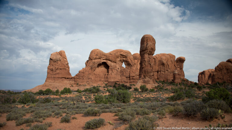 Small arch opening in a broad sandstone wall framed by tall fins and towers beneath cloudy skies.