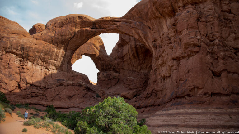 Large sandstone arch frames a smaller opening and a lone visitor near the rock wall.