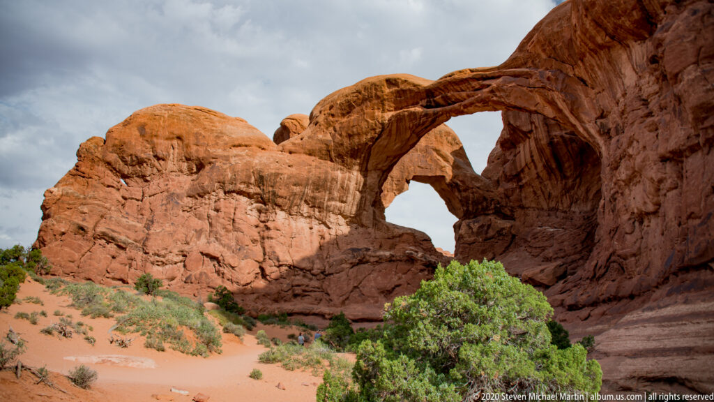 Intersecting sandstone spans rise above a sandy slope with green shrubs at the base in sunlight.