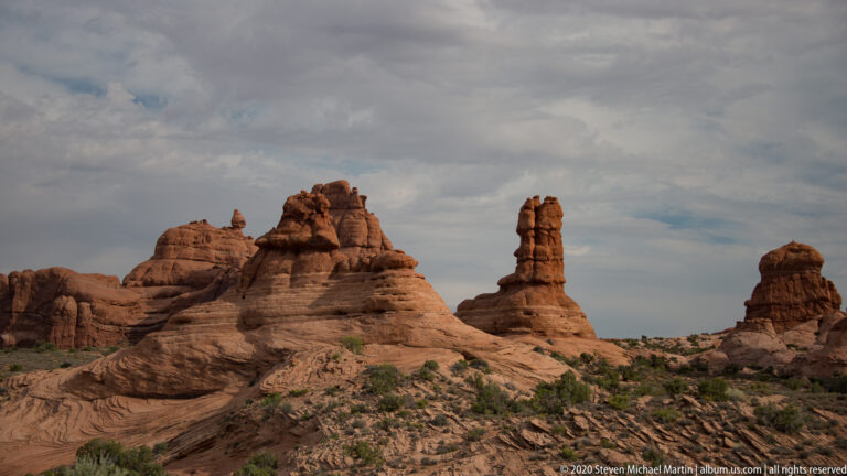 Several sandstone pinnacles rise separately along a scrub-covered slope under a cloudy sky.