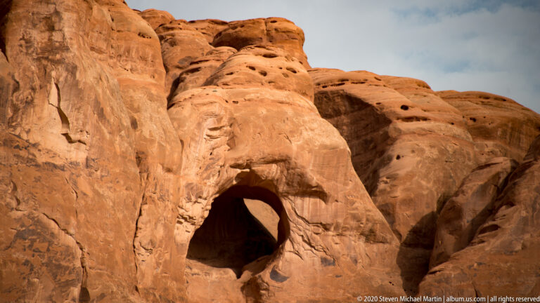 Deep dark cavity and smaller pits in a sunlit, weathered sandstone face.