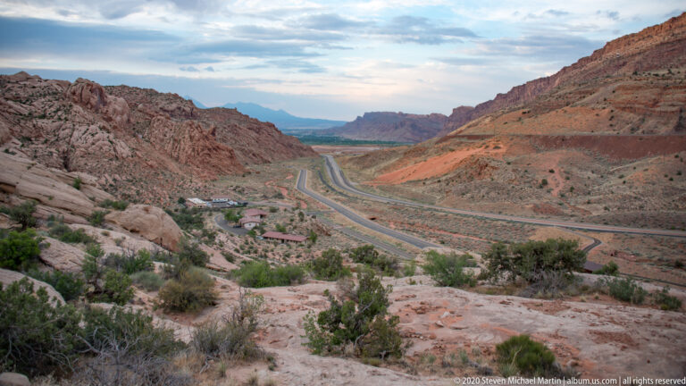 A highway winds down a broad desert valley between layered sandstone slopes.