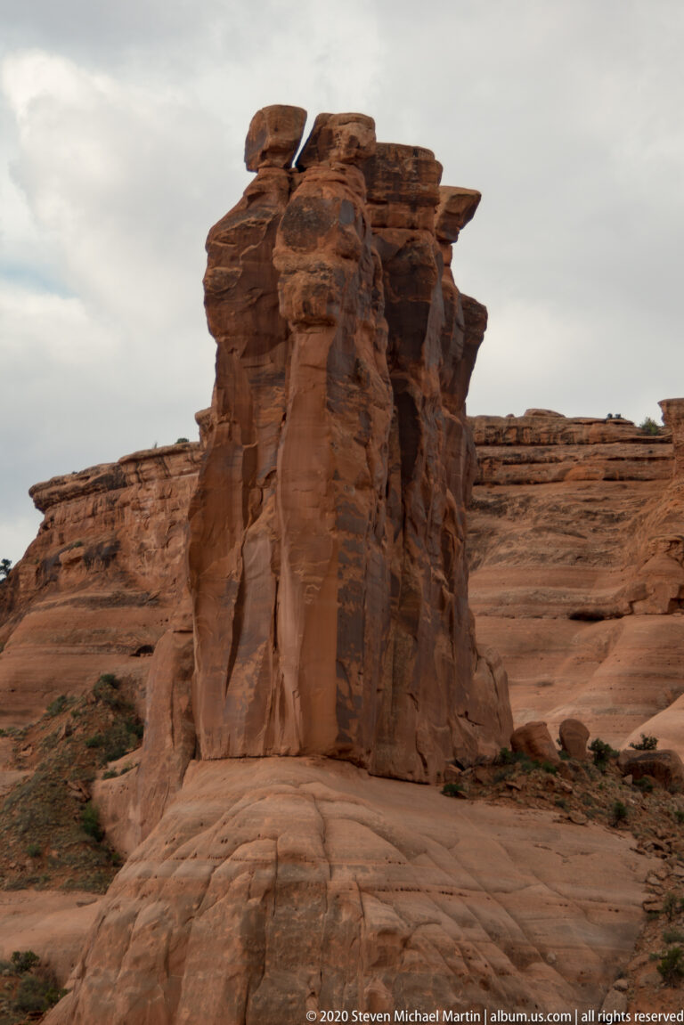 Tall fractured sandstone fin on a smooth bench, with layered cliffs behind it.
