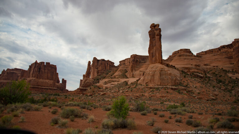 Narrow sandstone pillar rises from a rounded base before rugged red cliff walls.