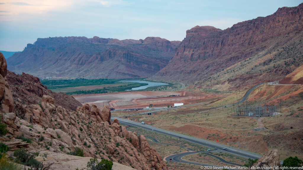 A highway curves toward a green river corridor in a wide valley framed by cliffs.