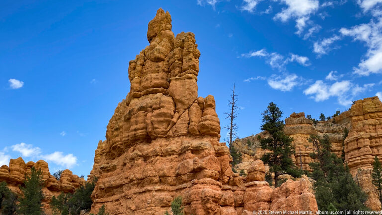 A tall banded hoodoo rises above smaller rock columns and pines beneath a blue sky.