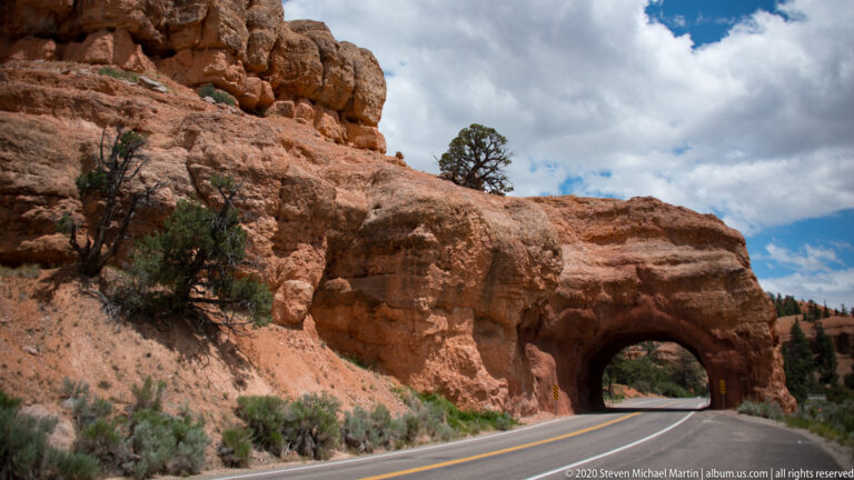 A two-lane road curves into a tunnel beneath a red sandstone overhang.