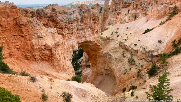 A broad natural opening pierces pale sandstone between orange cliffs and scattered pines.