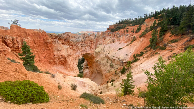 A natural arch sits in a pale cliff between orange rock and a forested slope.