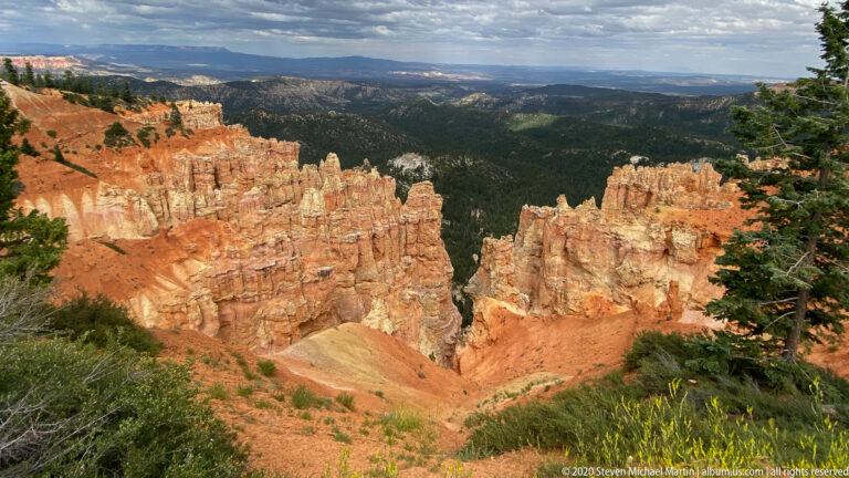 Orange hoodoos descend into a forested basin beneath a distant hazy plateau.