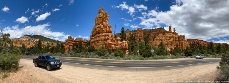 A pickup and curving road sit below red hoodoos and tall pinnacles beneath a clouded sky.