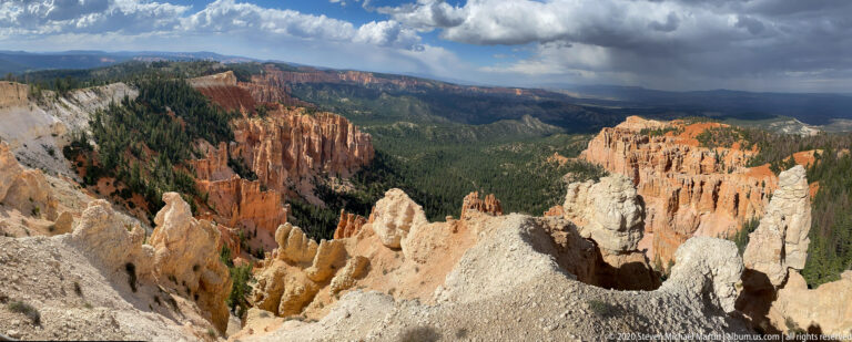 Pale hoodoos overlook an orange amphitheater and forest beneath a storm cloud.