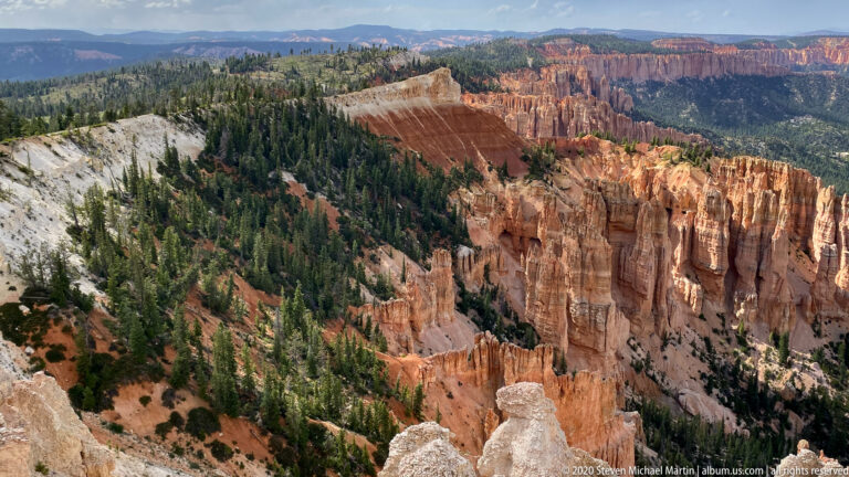 A long orange cliff edge descends into pines and hoodoos above a distant plateau.