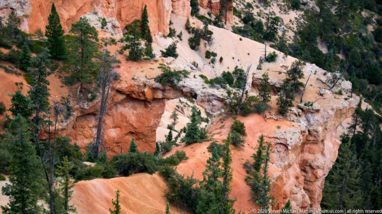 A broad opening pierces a pale ledge above orange slopes and scattered pines.