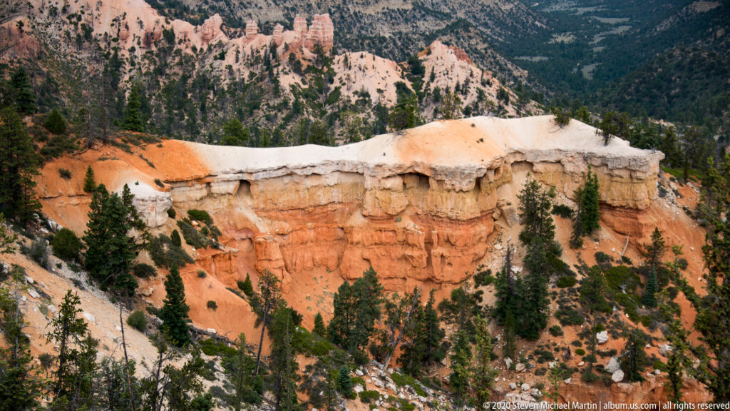 A pale caprock band tops an orange cliff above a forested slope.