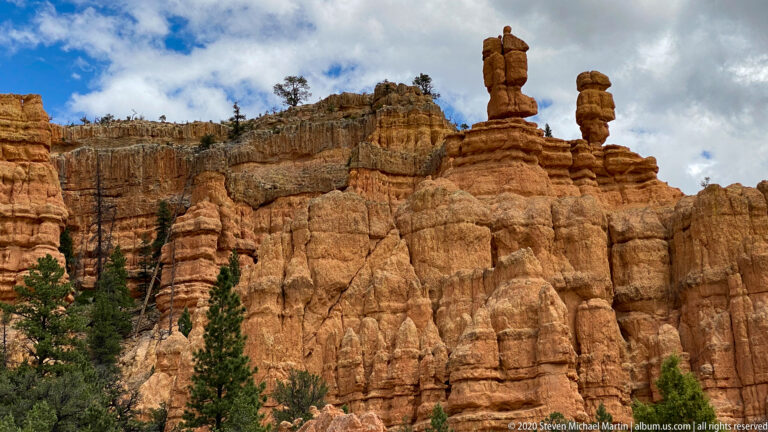 Two narrow hoodoo columns stand above a terraced sandstone wall dotted with pines.