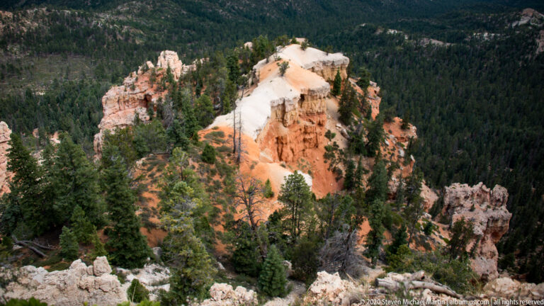 A pale-capped sandstone promontory rises above dense pines and nearby hoodoos below.