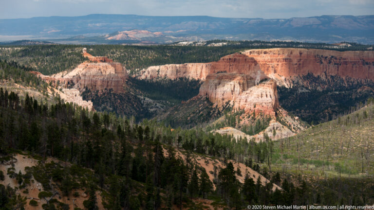 Orange and pale cliffs form a broad amphitheater amid dark forest, with a distant plateau under a cloudy sky.