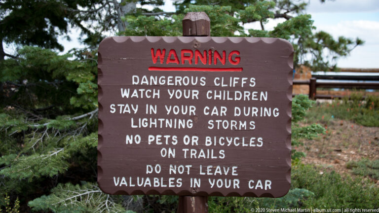 A brown sign lists warnings about cliffs, children, lightning, pets, bicycles, and valuables at a park overlook.