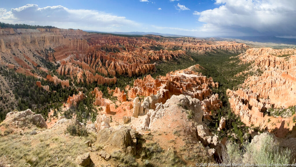 A broad panorama shows orange hoodoos descending into a pine-covered basin beneath distant cliffs and clouds.