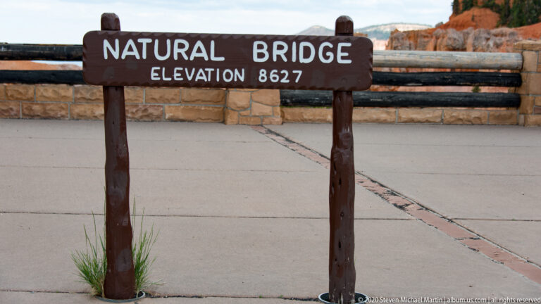A wooden Natural Bridge marker gives an elevation of 8,627 feet in a paved overlook area.