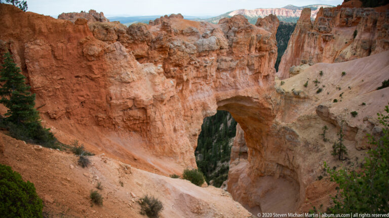 An orange natural arch spans a forested ravine between massive sandstone walls and hoodoo-covered cliffs.