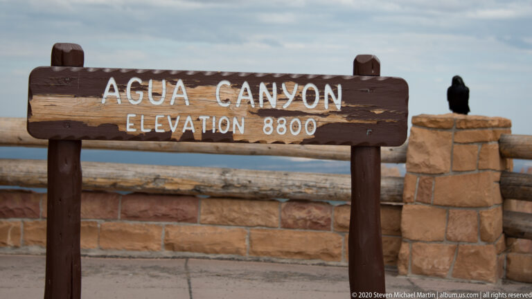 An Agua Canyon sign gives an elevation of 8,800 feet, with a black raven perched on a nearby stone pillar.