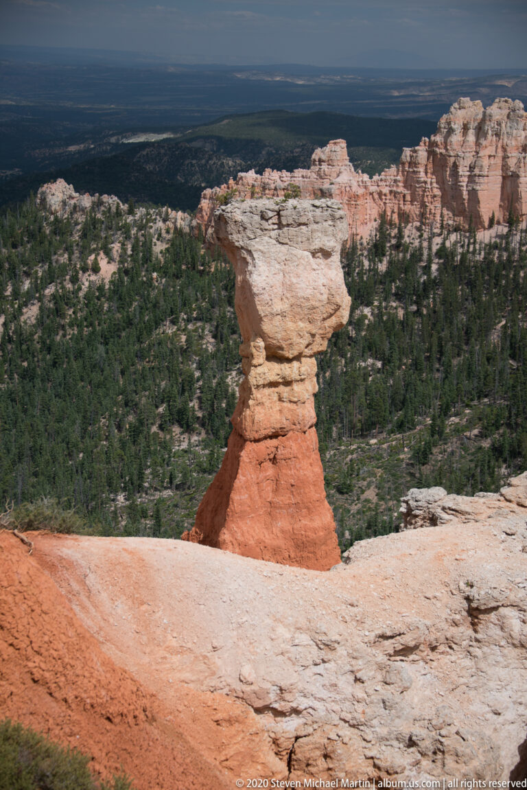 A pale caprock balances on a narrow orange pedestal above a forested plateau and canyon basin.