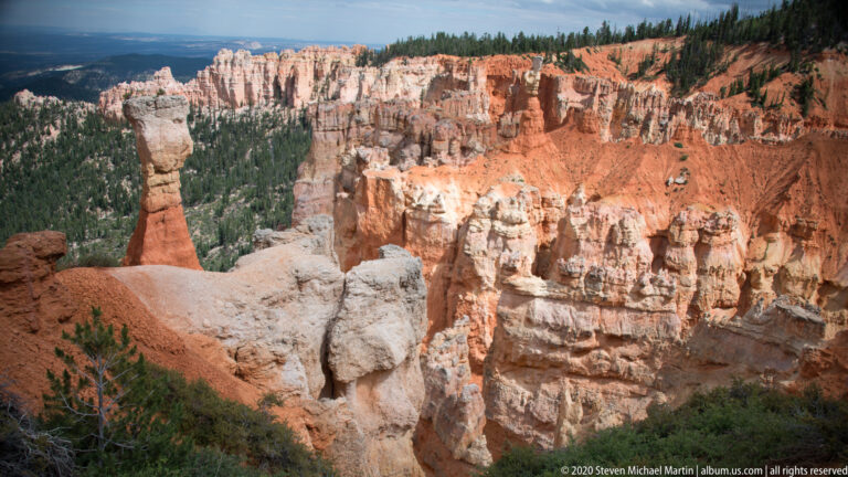 Pale hoodoos on a near ledge face an orange cliff wall across a forested canyon basin.