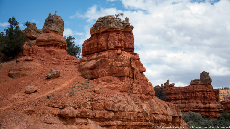 Layered sandstone pillars with irregular caps rise above a steep red slope.