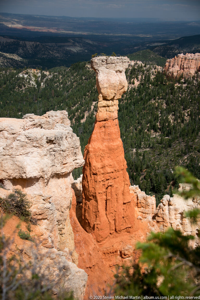 A broad pale capstone rests on an orange pedestal at the canyon rim above a forested plateau.