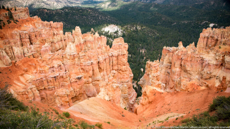 Orange hoodoos descend between steep canyon walls toward a forested basin and distant pale plateau.