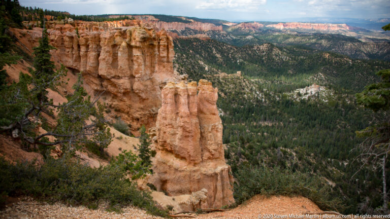 A tall orange hoodoo overlooks a forested valley and distant pale cliffs, with branches framing the left edge.