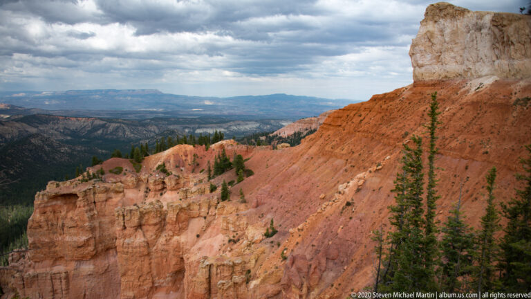 A pale cliff and orange slope overlook layered wooded ridges and a broad distant plateau beneath clouds.