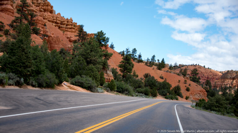 A marked highway curves beside pines and a red sandstone hillside under blue sky.