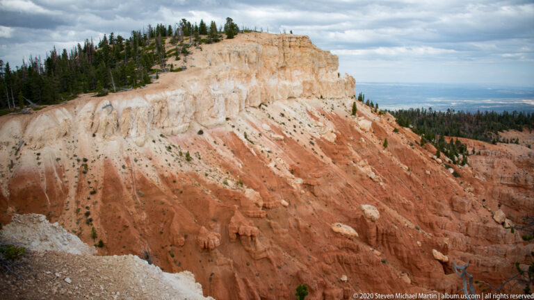 Cream and orange canyon cliffs fall from a pine-covered rim toward distant blue ridges.