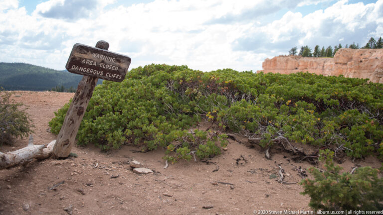 Warning sign for dangerous cliffs beside a sandy path and dense green shrubs.