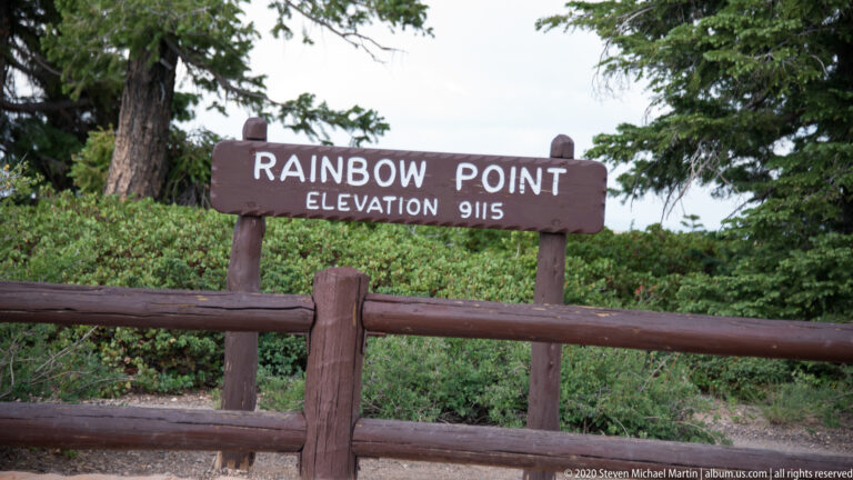 Rainbow Point sign gives an elevation of 9,115 feet above a wooden fence and shrubs.