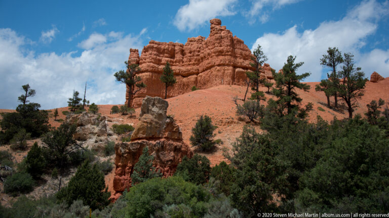 A small capped hoodoo stands before an orange cliff and scattered pines.