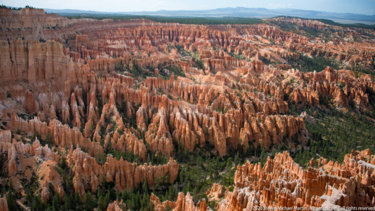 Expansive panorama of orange hoodoos and dark forest patches extending toward a distant plateau.