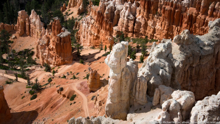 Pale rock spires stand above orange slopes and scattered green trees in the canyon.