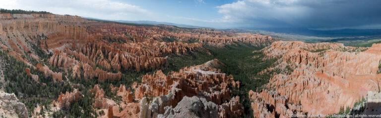 Orange hoodoos stretch toward a distant valley beneath a dark cloud and patches of sunlight.
