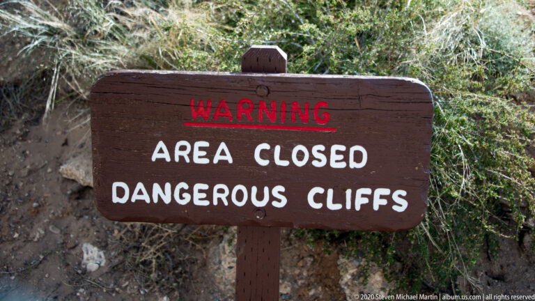Wooden warning sign says the area is closed because of dangerous cliffs, amid shrubs and rock.