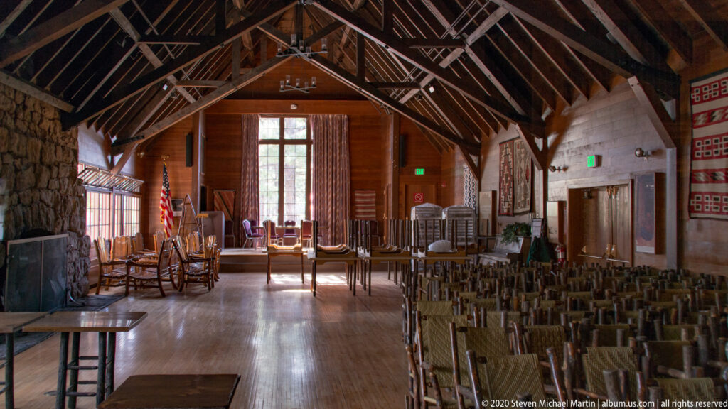 Rustic timber room with exposed roof trusses, stone walls, tables, and rows of chairs.