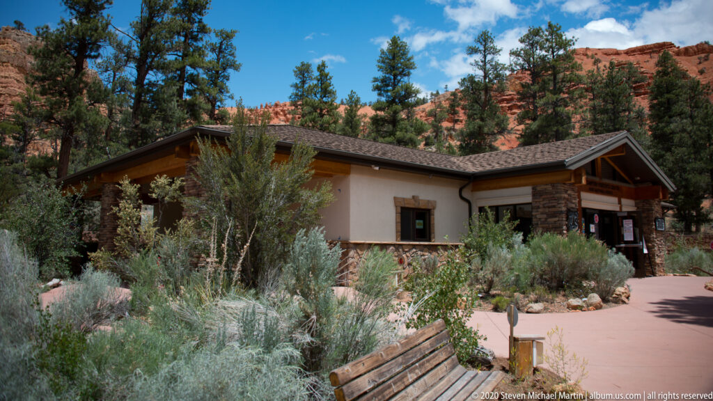 A low visitor center with a sloped roof stands among shrubs and pines beneath red cliffs.