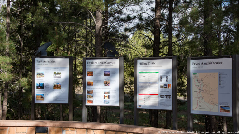 Four visitor panels about park amenities, Bryce Canyon, hiking trails, and Bryce Amphitheater stand beneath evergreen trees.