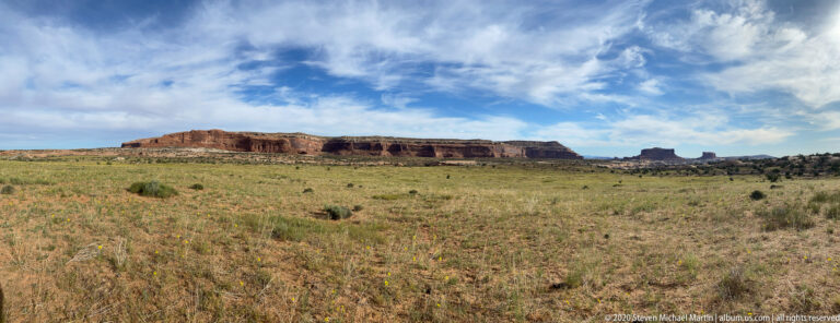 Grassy plateau stretches toward a long red mesa under broken clouds.